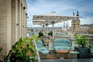 A rooftop terrace with outdoor furniture, a large umbrella, and a view of the cityscape including a historic building. - Motel One Koblenz (Koblenz)