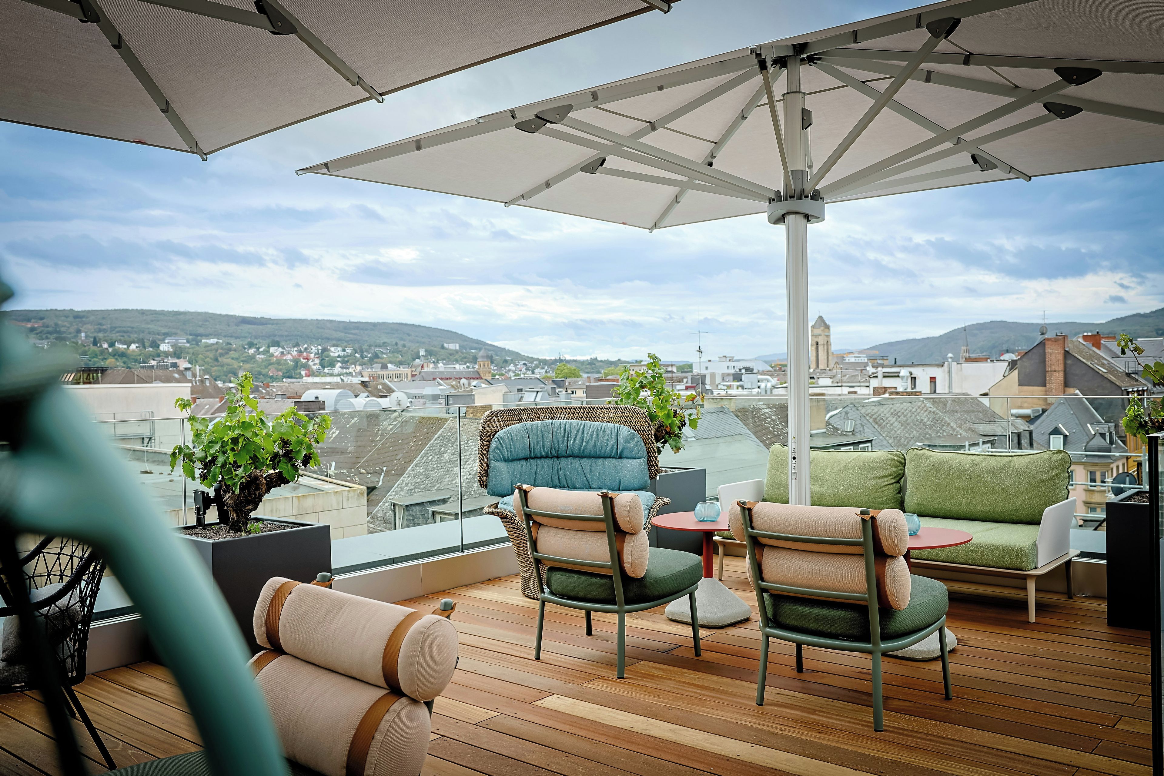Un balcón con vista a un paisaje urbano, amoblado con sillas y una mesa, y una sombrilla grande que proporciona sombra.