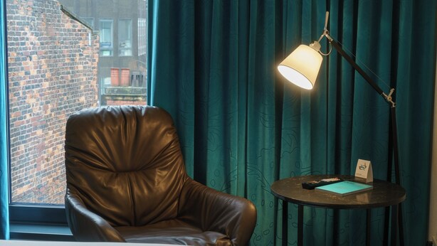 A leather armchair by a window with a view of a brick building, a desk with a lamp, and a small table with a card and remote control. - Motel One Koblenz (Koblenz)