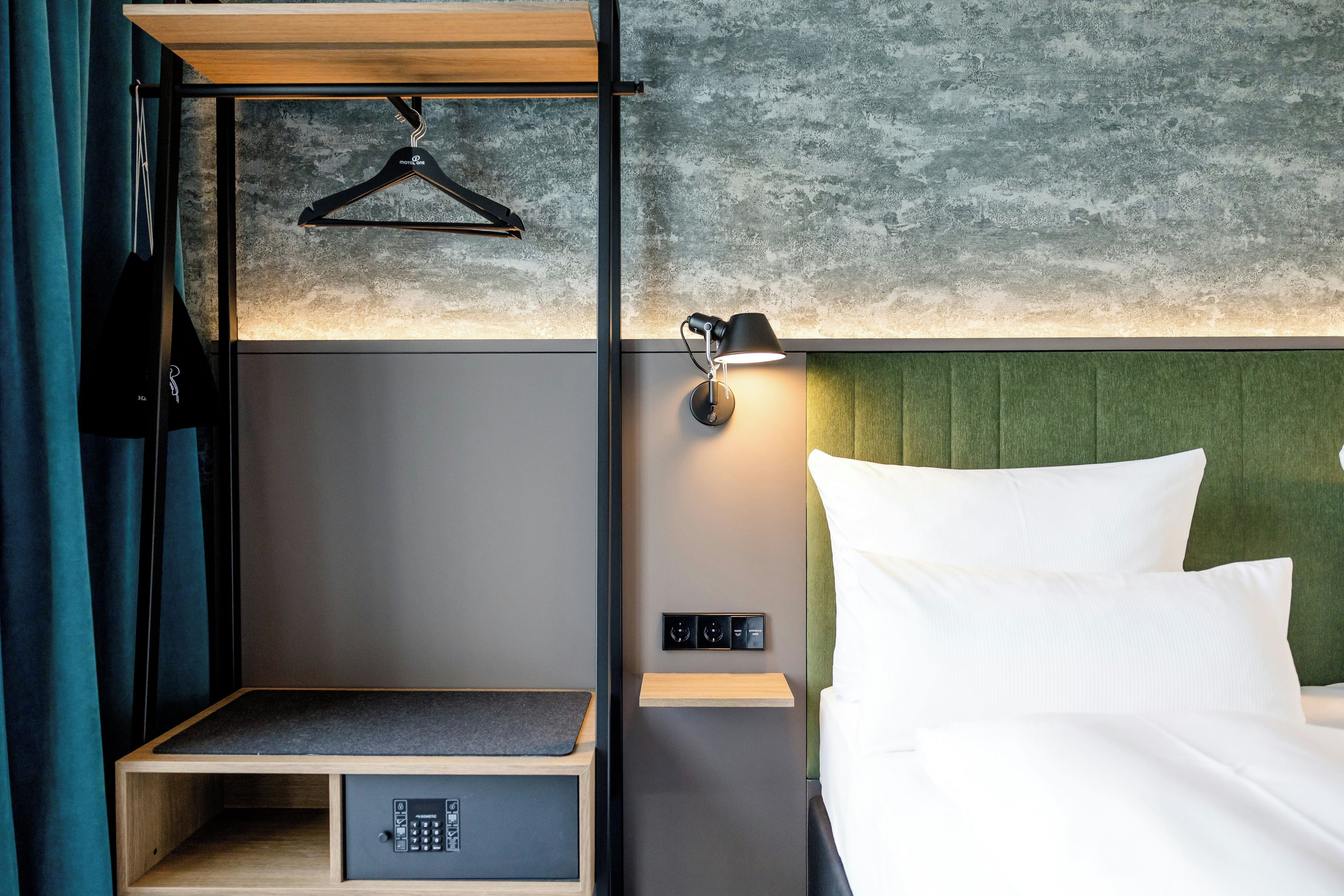 Ein Hotelzimmer mit einem Bett, einem Nachttisch, einer Hängelampe, einer Steckdose und einem Kleiderhaken.