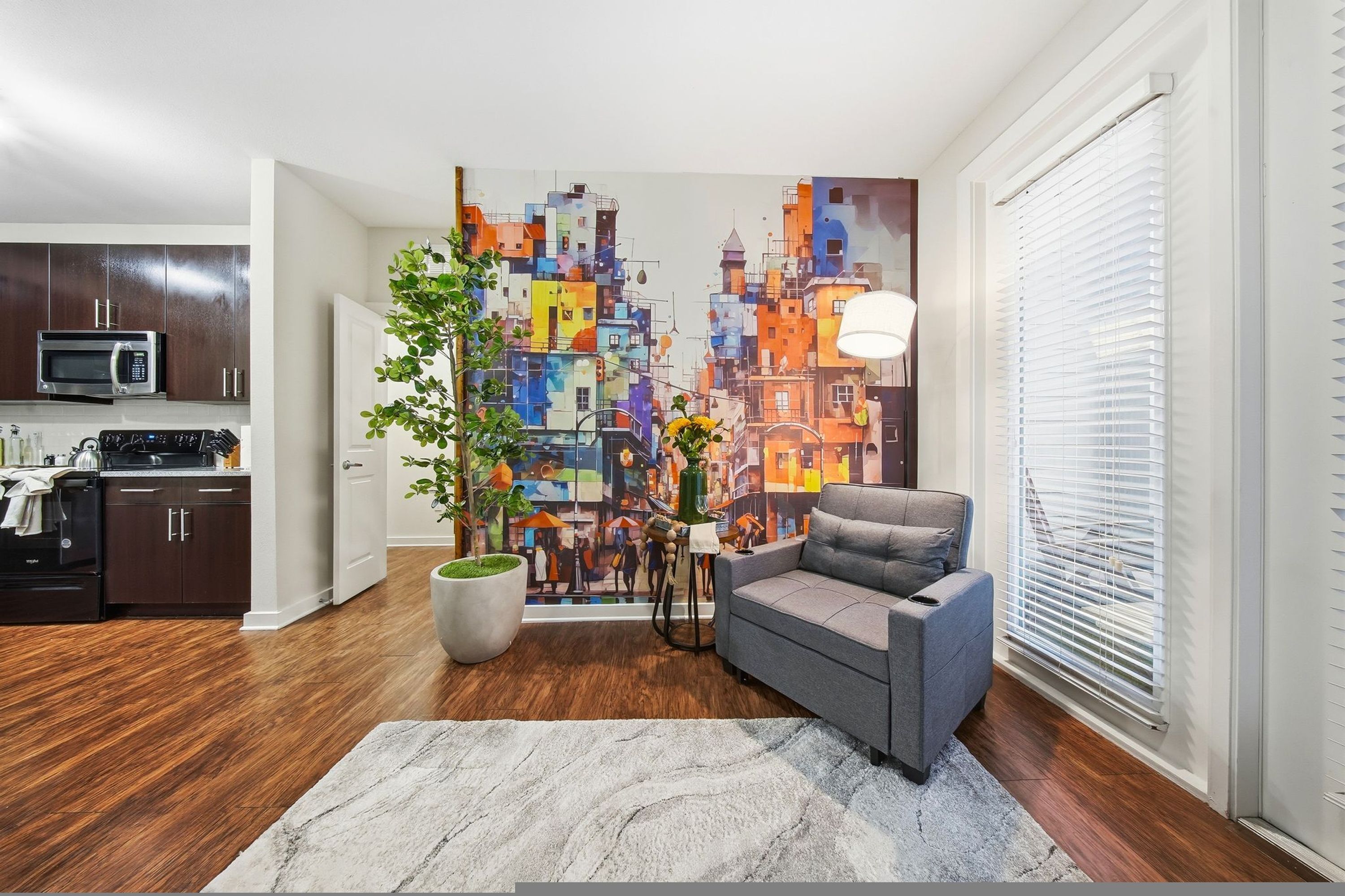 A modern living room with a gray sofa, a potted plant, a colorful cityscape mural, and a kitchen in the background.