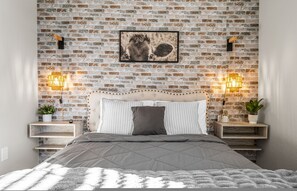 Une chambre avec un mur décoré d’un motif en briques, un tableau encadré, des tables de chevet avec des lampes et un lit recouvert d’une couette grise.