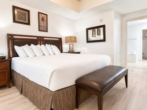 Quarto de hotel com uma cama grande, uma mesinha de cabeceira, uma luminária, um banco e um espelho na parede.