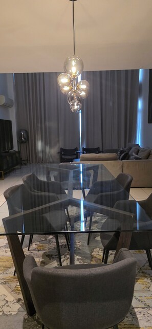 A modern dining area with a glass table and chairs, a hanging light fixture, and a sofa in the background. - ebs properties ghana (Accra)