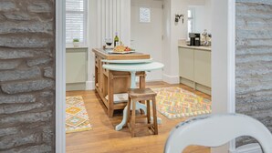 Een keuken met een ronde tafel, houten stoelen, een raam met jaloezieën, een deur en een klok aan de muur.