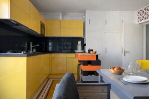 A compact kitchen with yellow cabinets, a black stove, and a dining area with a table set for two. - Beautiful family home for 8 guests – Les Roses (Les Sables-d´Olonne)