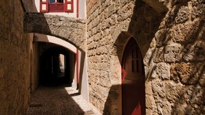 A narrow stone alleyway with an arched stone structure, a cobblestone path, and a building with a red door and window shutters. - Vivila in the medieval city of Rhodes (Rodos)