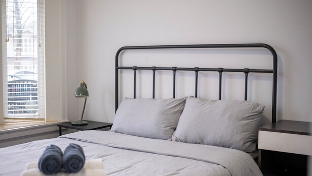 Un lit bien fait, avec des oreillers, une lampe et une fenêtre équipée de stores.