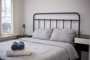 Ein ordentlich bezogenes Bett mit Kissen, einer Lampe und einem Fenster mit Jalousien.