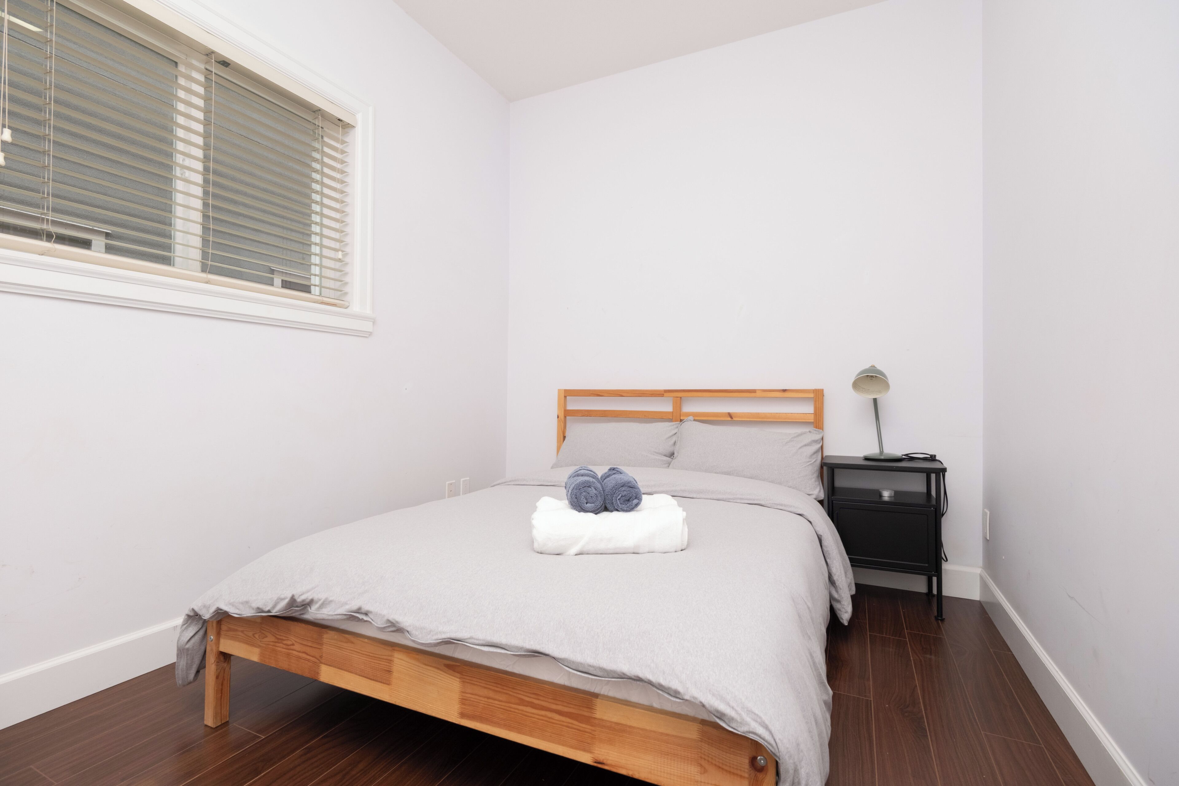 A neatly made bed with a wooden headboard, a bedside table with a lamp, and a window with blinds.
