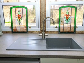 A kitchen with a stainless steel sink, two windows with stained glass, and a view of a brick wall and greenery outside. - Milburn Cottage (Keighley)