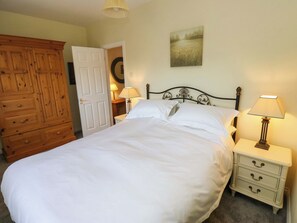 A neatly made bed with white linens, a wooden wardrobe, a bedside table with a lamp, and a wall-mounted painting. - Milburn Cottage (Keighley)