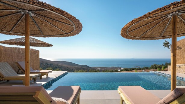 Ein Poolbereich mit Liegestühlen und Reedschatten, mit Blick auf das klare, blaue Meer und die entfernten Hügel.