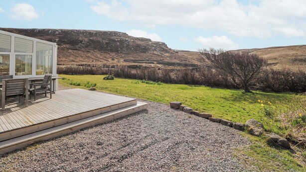 Un bâtiment moderne doté de grandes fenêtres, d’une terrasse en bois et d’un chemin de gravier menant à une zone herbeuse d’où l’on peut admirer une colline.