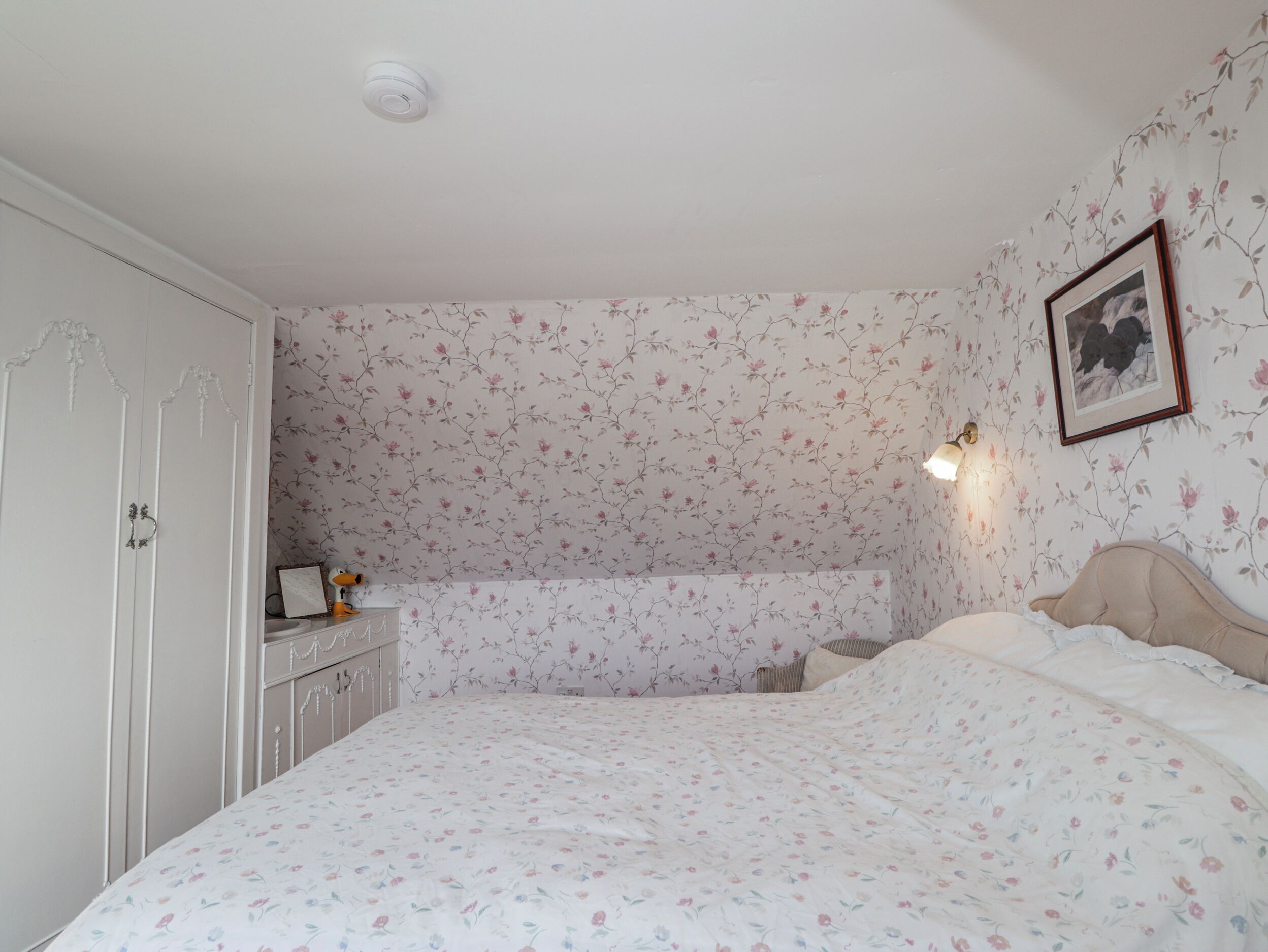 A bedroom with a floral wallpaper, a bed with a floral bedspread, a wardrobe, a bedside table with a lamp, and a framed picture on the wall.