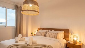 A hotel room with a large bed, a wooden headboard, a hanging light fixture, a bedside table with a lamp, and a window with curtains. - Casares Rooftop Apartment by Costarentals (Casares)