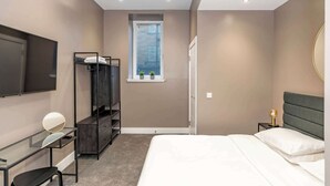 Een slaapkamer met een bed, een bureau, een televisie, een kledingkast, een raam met zonwering en een bedlampje.