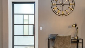 Een kamer met een grote wandklok, een bureau met stoel en een deur naar een balkon.
