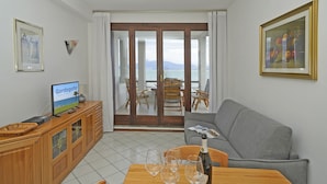 Ein Wohnzimmer mit Fernseher, einem Holzschrank, einem Esstisch mit Weingläsern, einer Couch und Blick durch eine Glastür nach draußen.