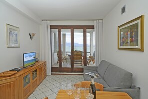 Ein Wohnzimmer mit Fernseher, einem Holzschrank, einem Esstisch mit Weingläsern, einer Couch und Blick durch eine Glastür nach draußen.