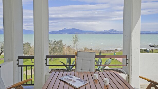 Ein Holztisch mit einer weißen Tasse und einer Speisekarte, eine Bank und Blick auf einen See mit Bergen im Hintergrund.