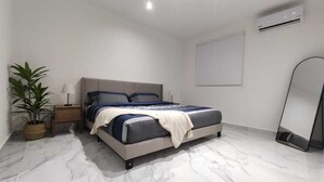 A bedroom with a bed, bedside table, lamp, potted plant, air conditioning unit, and a mirror. - Ground Floor Apartment in Ombú Residential Cancun (Cancún)