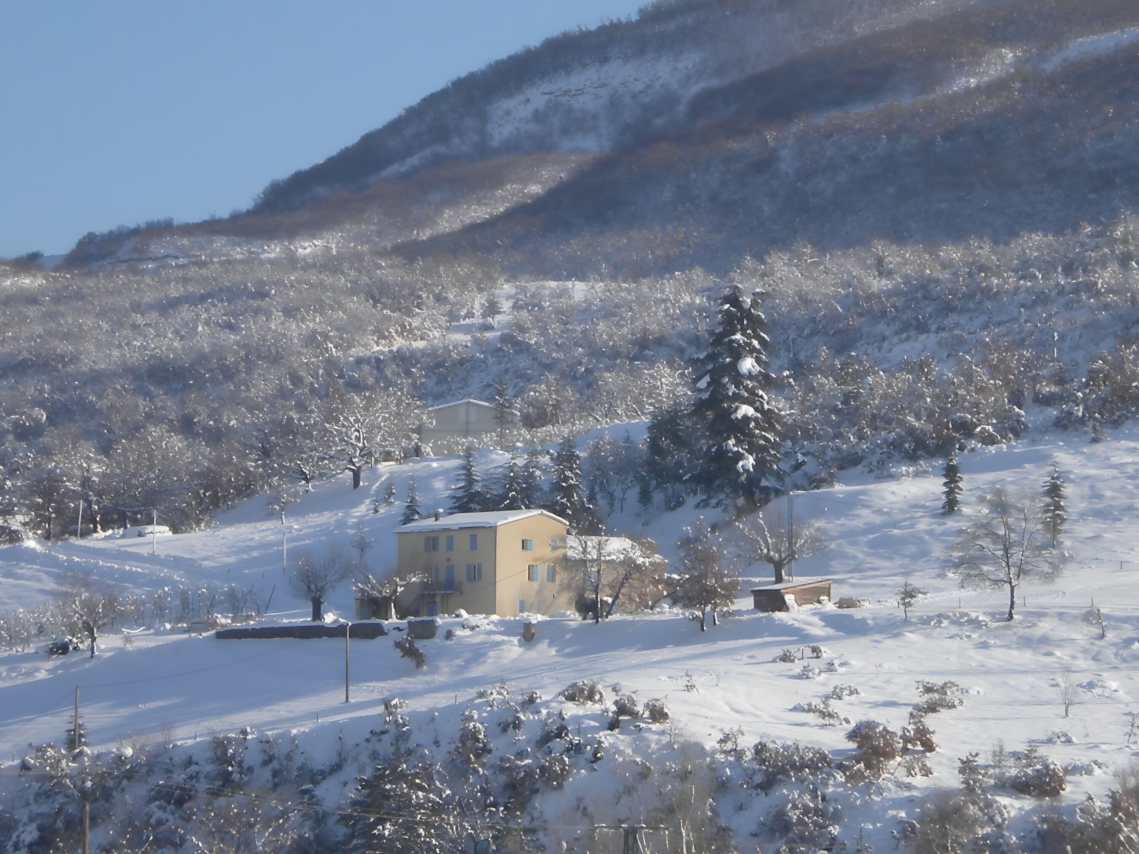A snow-covered landscape with a building, trees, and a hill.