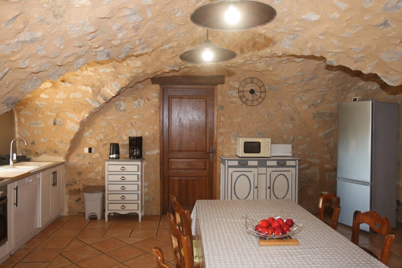 Una cocina con paredes de piedra, una puerta de madera, un microondas, un refrigerador, un lavamanos y una mesa de comedor con sillas.