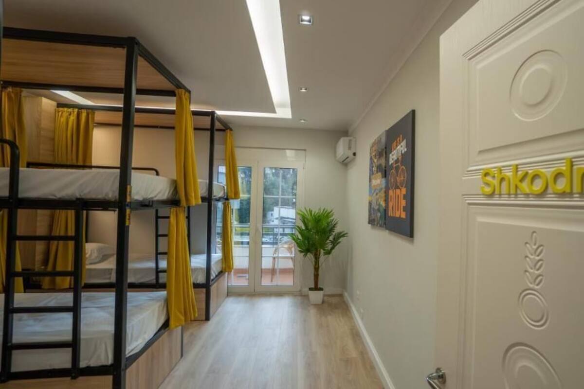 A hotel corridor with bunk beds, a potted plant, and a door with the name 'Shkodra' on it.