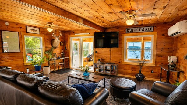 L'intérieur d'un chalet en rondins, avec un canapé en cuir, un téléviseur, une enseigne indiquant « APPÂTS ET MATÉRIEL DE PÊCHE », et une vue sur l'extérieur à travers de grandes fenêtres.