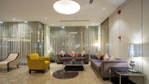 A modern hotel lobby with a sofa set, a yellow armchair, a glass coffee table, and a side table with a lamp. - Topaz Hotel Suites (Riyadh)