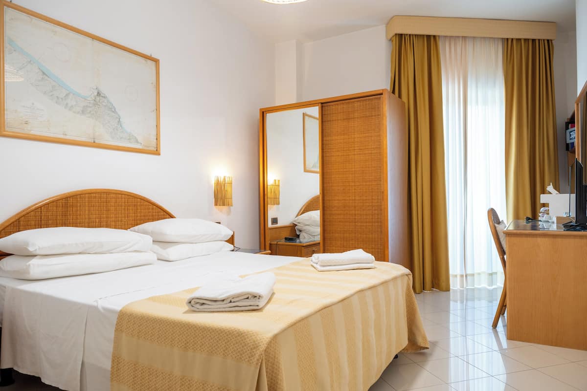 A hotel room with a bed, a desk, a chair, a mirror, and a large window with curtains.
