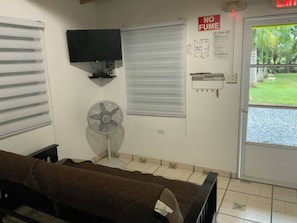 Une chambre avec un lit, un téléviseur fixé au mur, un ventilateur, une fenêtre avec des stores, une porte sur laquelle est apposée une pancarte indiquant « INTERDIT DE FUMER », et une vue sur un jardin.