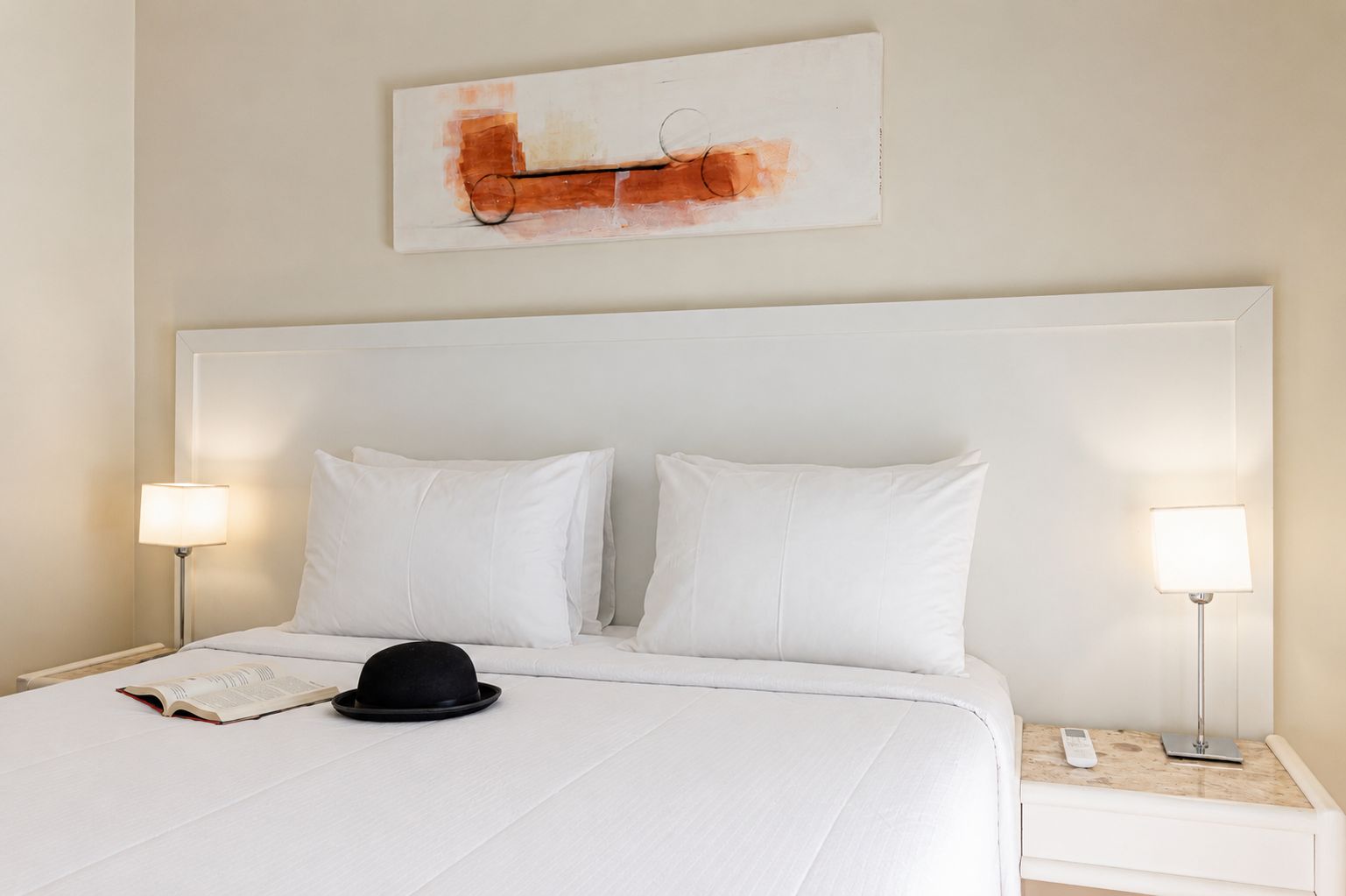 Una habitación de hotel con una cama grande, dos lámparas de noche, un libro y un sombrero sobre la cama.