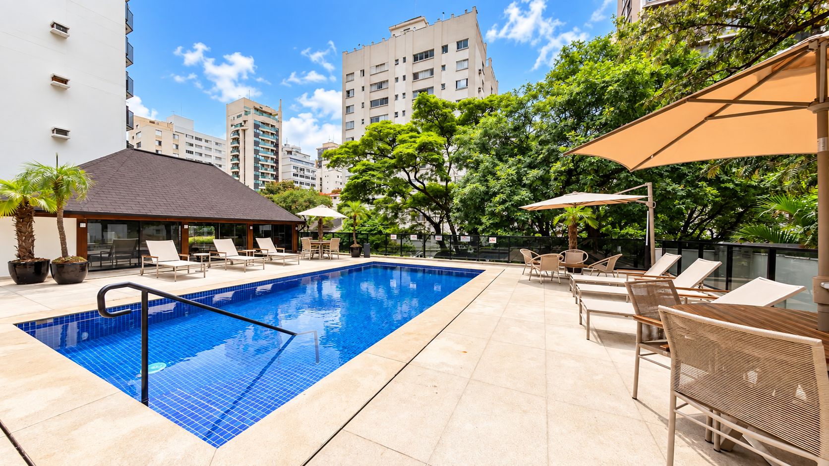 Piscina com espreguiçadeiras e guarda-sóis, cercada por vegetação e edifícios.