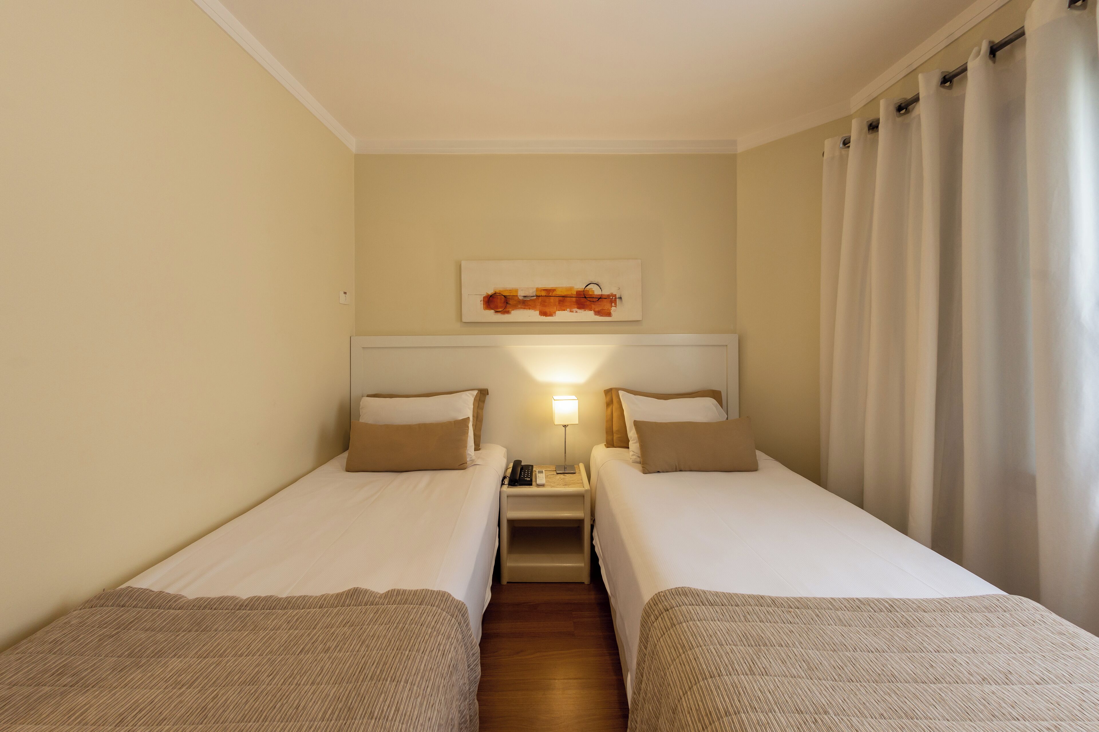 Een hotelkamer met twee enkele bedden, een nachtkastje met een lamp, een schilderij aan de muur en een raam met gordijnen.
