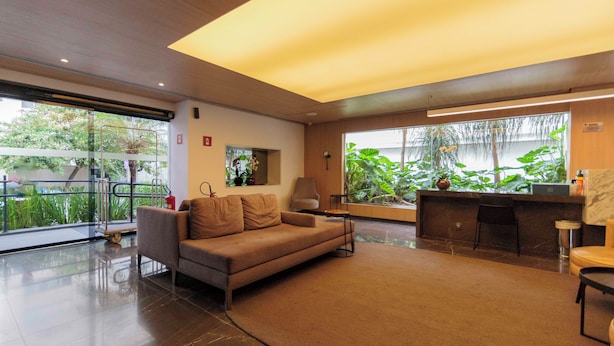 A modern interior with a sofa, a small table, a lamp, a bar area, and large windows with plants. - Charlie Victoria Itaim (São Paulo)