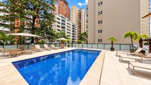 Una piscina con sillones de playa y sombrillas, rodeada de edificios altos.