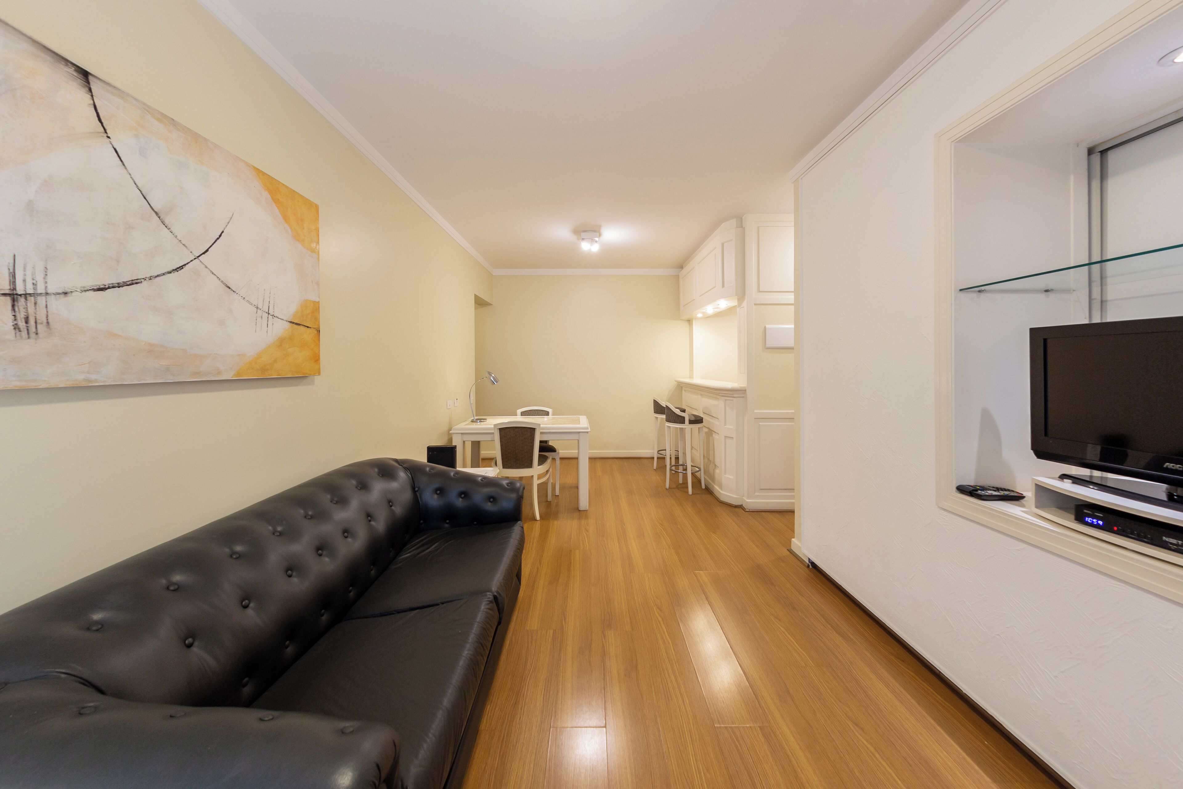 A modern living room with a black leather sofa, a dining table with chairs, a kitchen area with cabinets, and a flat-screen TV mounted on the wall.