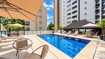 Piscina con sillas de descanso y sombrillas, rodeada de edificios.
