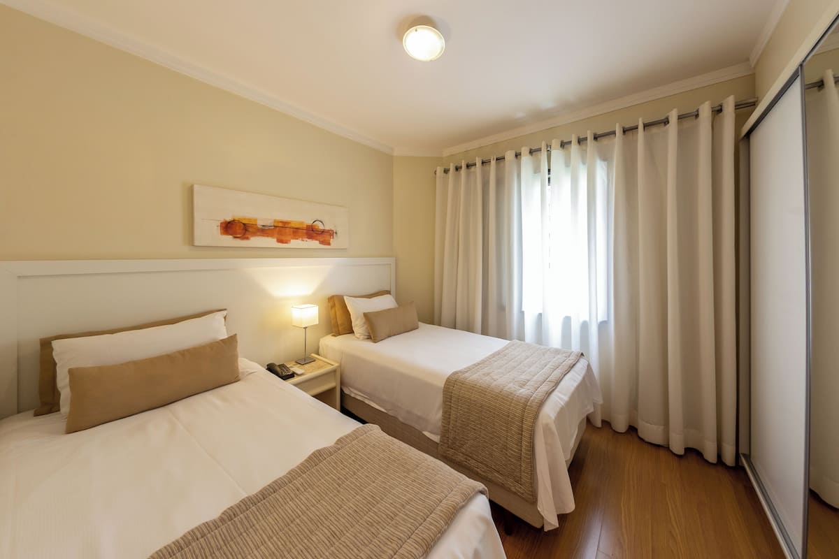 Une chambre d’hôtel avec deux lits, une table de chevet avec une lampe, un tableau au mur et une fenêtre avec des rideaux.