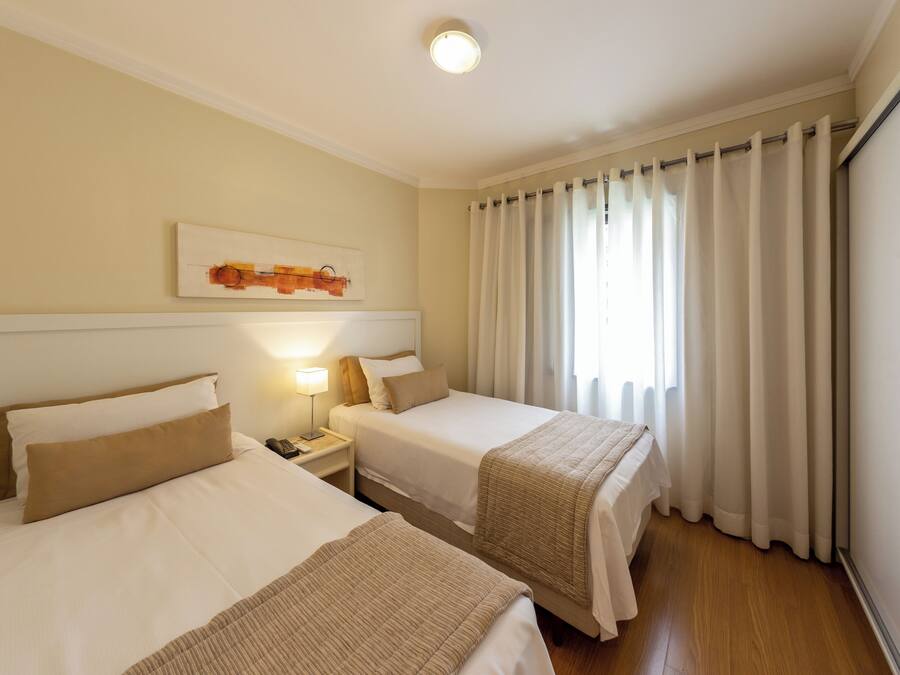 A hotel room with two beds, a nightstand with a lamp, a painting on the wall, and a window with curtains.