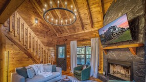A wooden cabin interior with a staircase, a fireplace, a sofa, a chair, and a television mounted on a stone wall. - Moon Lake Retreat - Kayaks, Fishing, Private shoreline, Hot tub, Room to Roam (Blue Ridge)