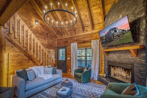 A wooden cabin interior with a staircase, a fireplace, a sofa, a chair, and a television mounted on a stone wall. - Moon Lake Retreat - Kayaks, Fishing, Private shoreline, Hot tub, Room to Roam (Blue Ridge)