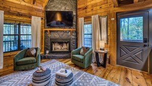 A log cabin interior with a stone fireplace, two green armchairs, a round coffee table, and a large TV screen. - Moon Lake Retreat - Kayaks, Fishing, Private shoreline, Hot tub, Room to Roam (Blue Ridge)