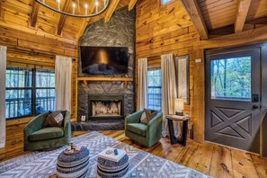 A log cabin interior with a stone fireplace, two green armchairs, a round coffee table, and a large TV screen. - Moon Lake Retreat - Kayaks, Fishing, Private shoreline, Hot tub, Room to Roam (Blue Ridge)