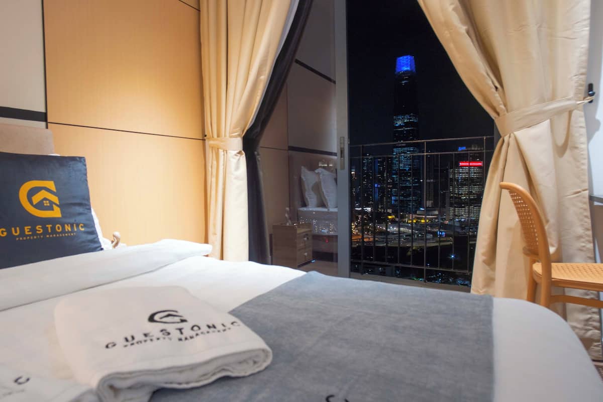 Habitación de hotel con cama, almohada con logotipo, toalla, silla y vista a un paisaje urbano de noche.
