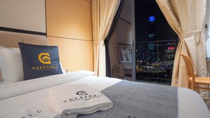 A hotel room with a bed, a pillow with a logo, a towel, a chair, and a view of a cityscape at night.