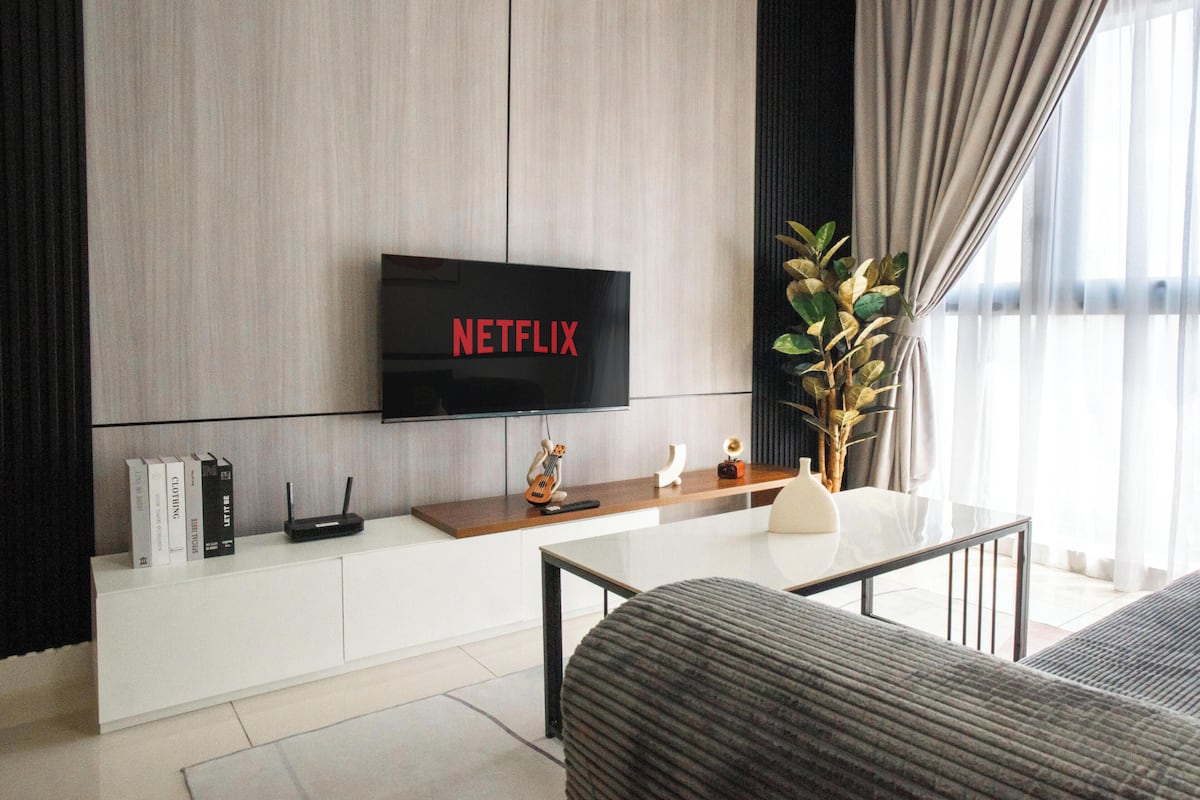 Un salón moderno con un televisor de pantalla plana mostrando 'NETFLIX', una consola blanca con libros, una planta pequeña y objetos decorativos.