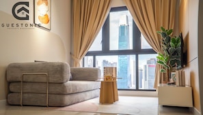 A modern living room with a sofa, a small wooden table, a potted plant, and a view of the cityscape through large windows.